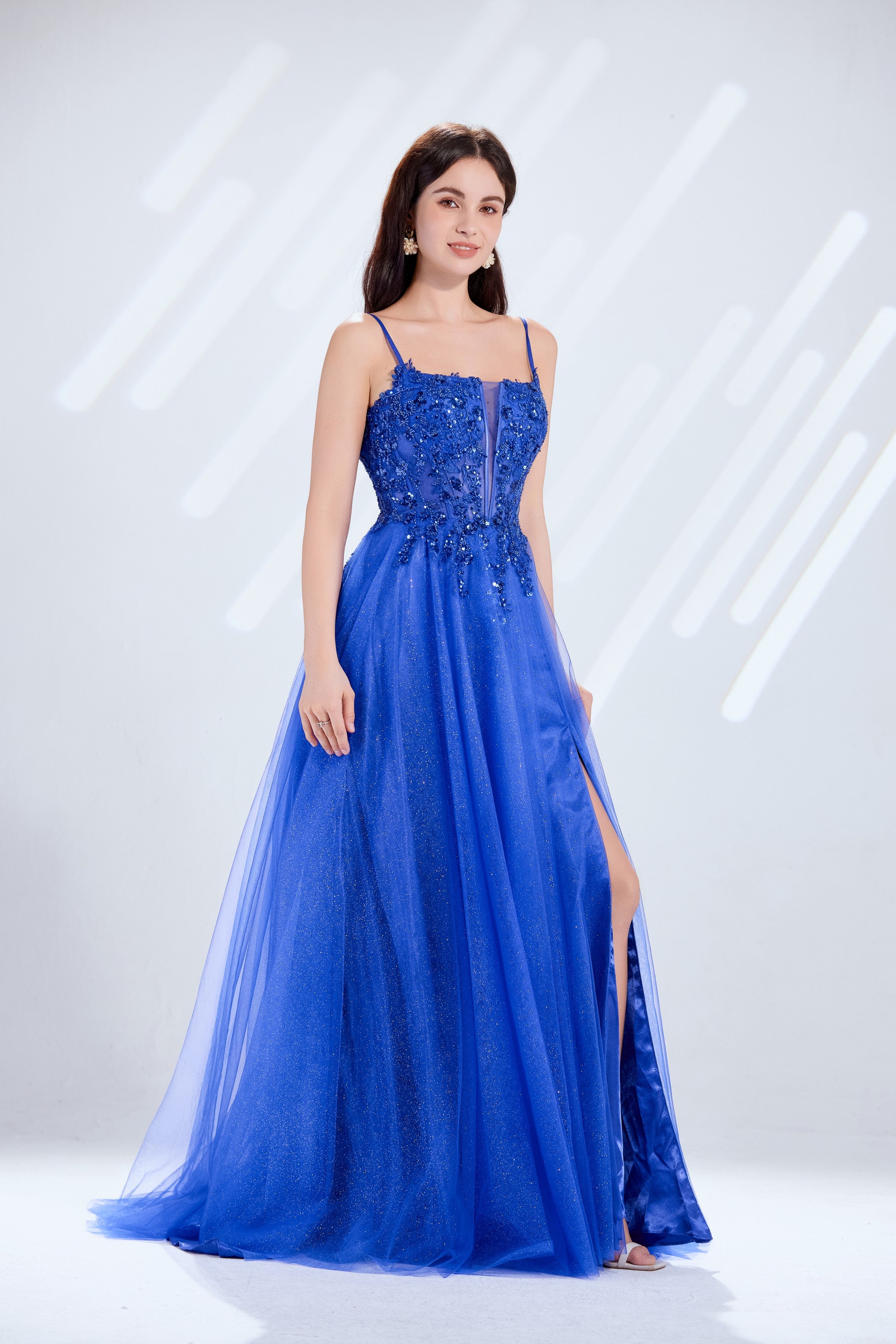 Model wearing a shimmering royal blue floor-length prom dress with a sequined lace bodice, spaghetti straps, and a high leg slit on a flowy glitter tulle skirt.