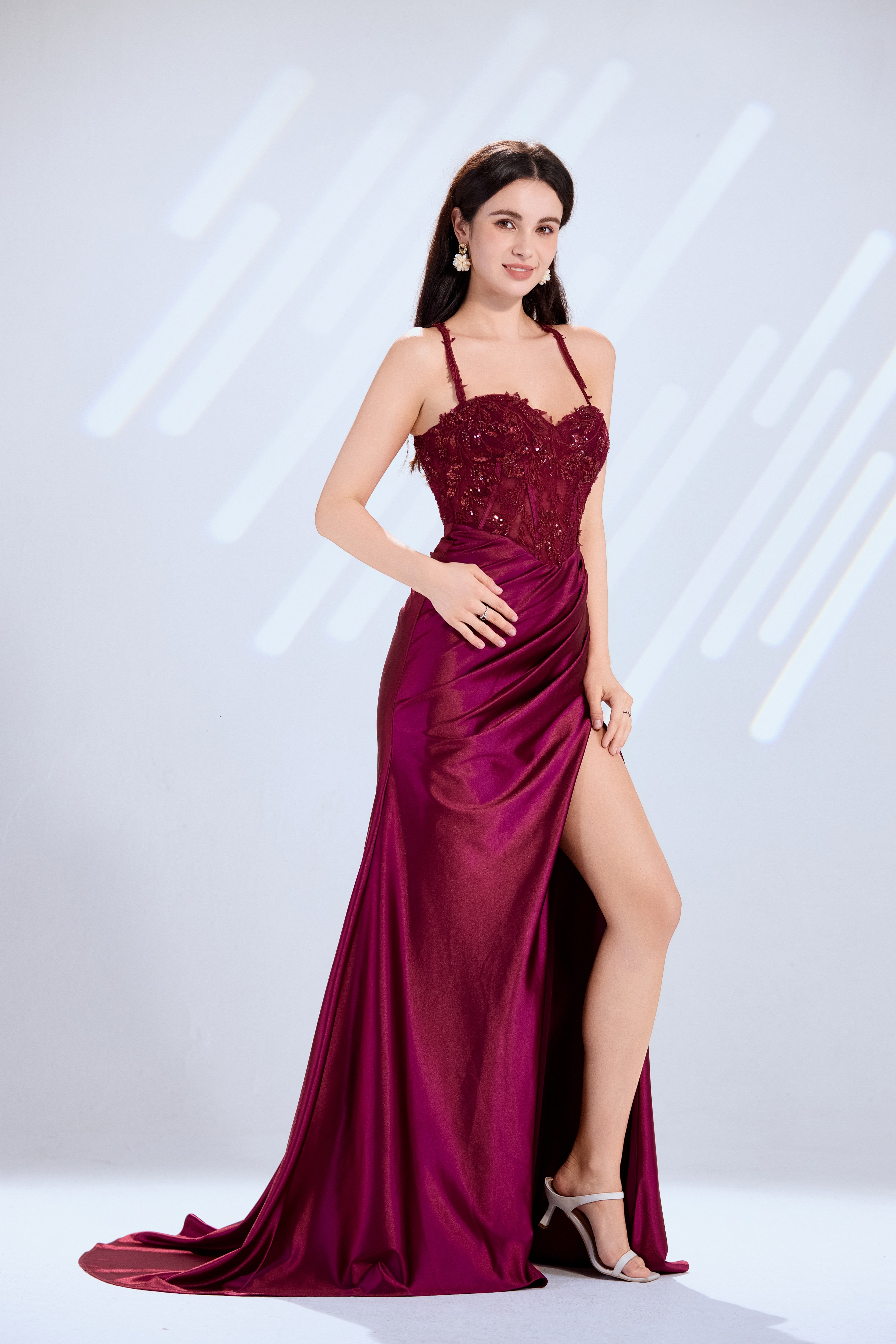 Model wearing a stunning burgundy evening gown with a sequined lace corset bodice, spaghetti straps, and a draped satin skirt featuring a high leg slit.