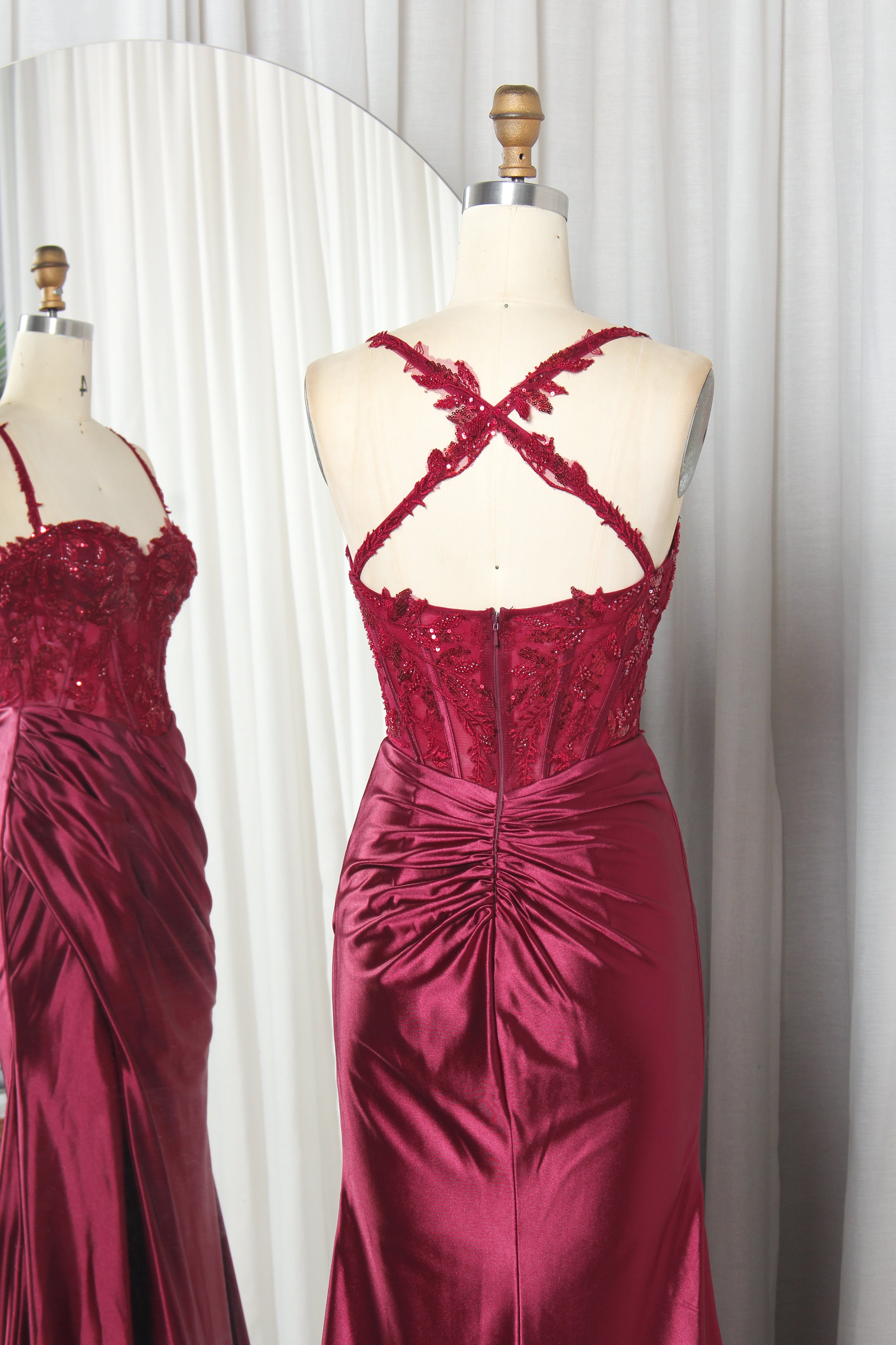 Model wearing a stunning burgundy evening gown with a sequined lace corset bodice, spaghetti straps, and a draped satin skirt featuring a high leg slit.