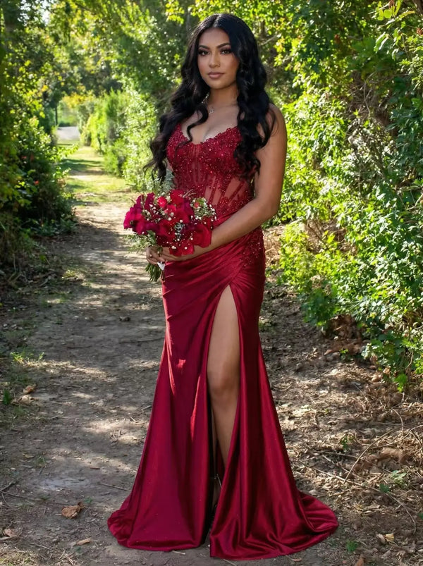 A beautiful woman posing in a burgundy red beaded corset gown with a thigh-high slit, standing on a rustic garden path, formal red carpet evening dress, perfect inspiration for prom 2026 or gala events.