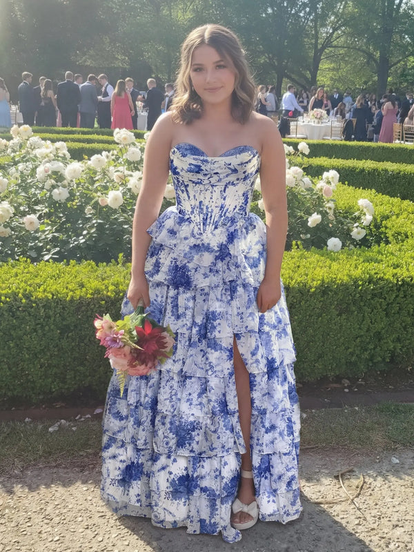A young woman wearing a stunning blue and white floral print corset gown with a tiered ruffle skirt and high side slit, posing in a sunny garden, formal prom dress inspiration, unique blue toile print evening gown for special occasions.
